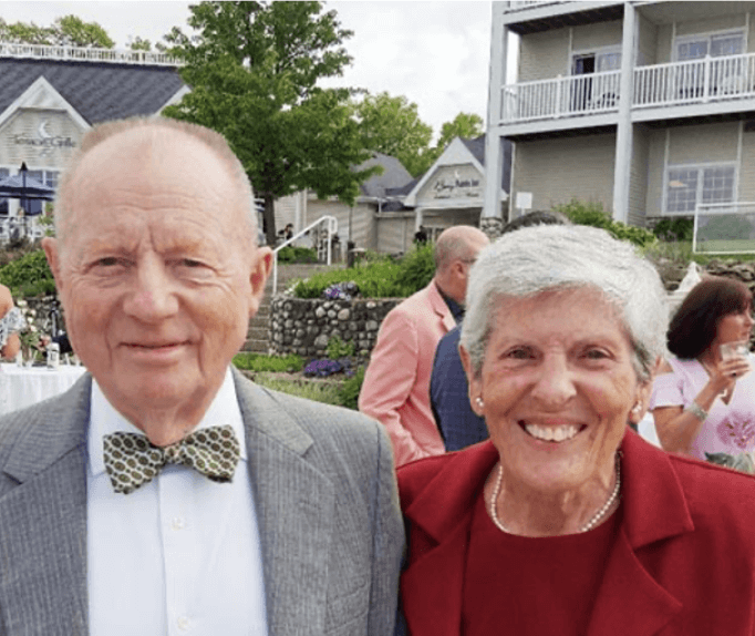 Smiling senior couple dressed in formal attire attending an outdoor event, with a scenic backdrop of greenery, flowers, and charming buildings.