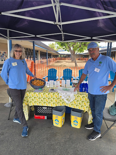 Two Senior Gleaners volunteers wearing blue shirts stand under a canopy in front of fresh produce, recycling bins, and eco-friendly goods.