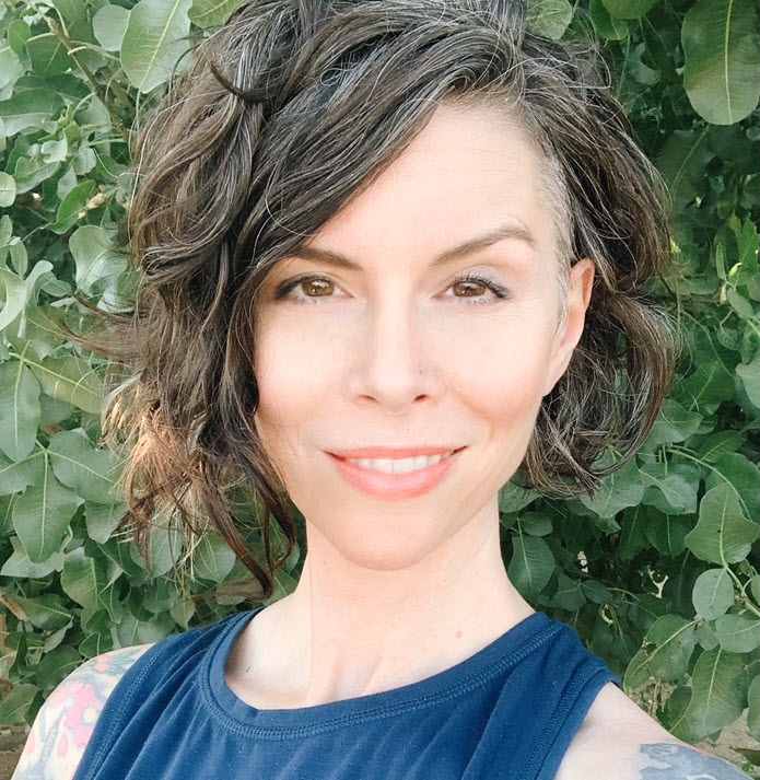 A woman with visible tattoos, dressed in a blue top, smiling at the camera.