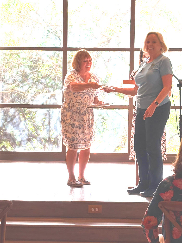 Two women on a raised platform during a grant recognition moment, with one presenting an item to the other.