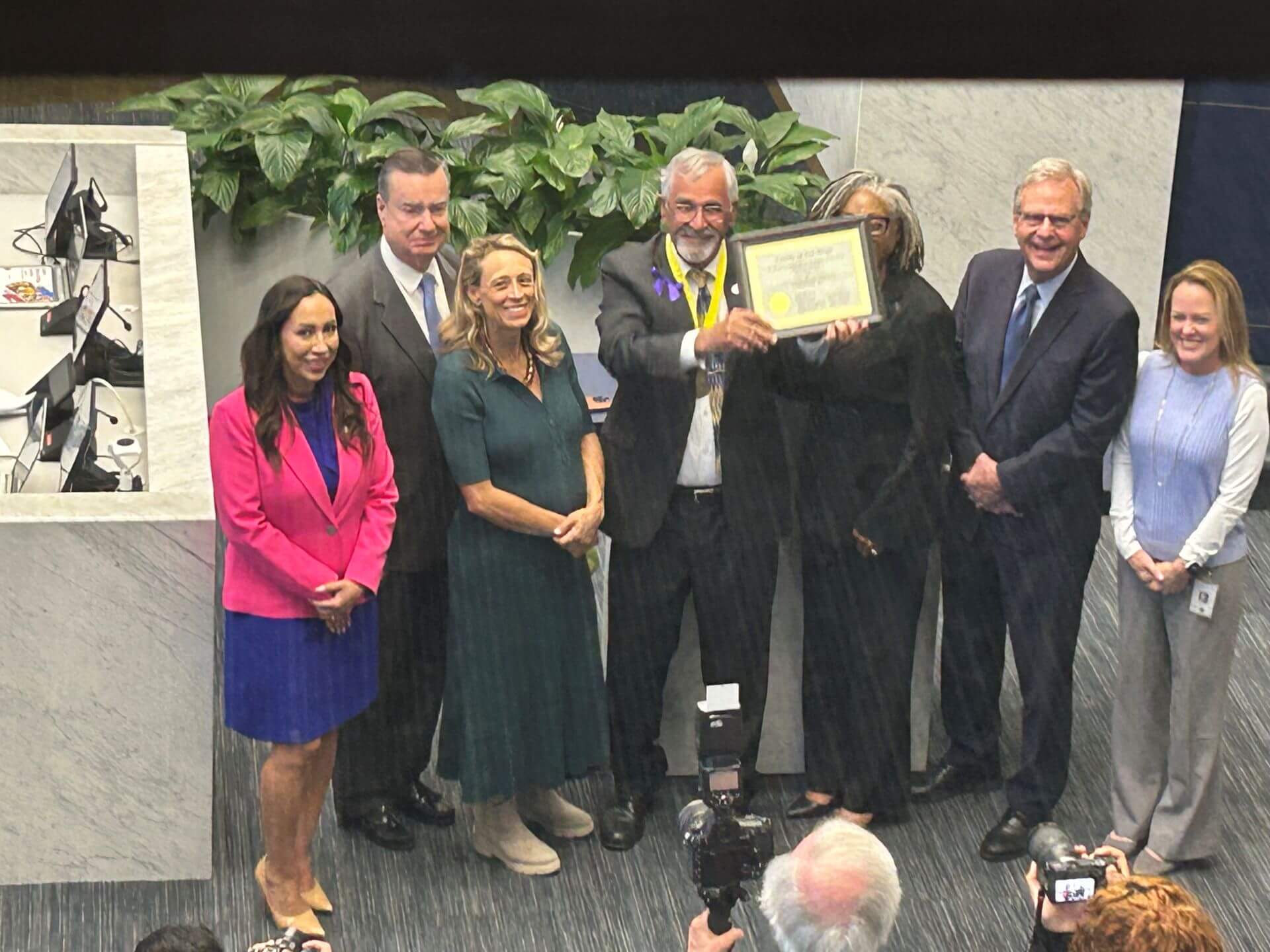 A group of seven people, some smiling, stand indoors as one person in the center holds up a framed certificate. There are plants and office equipment in the background.