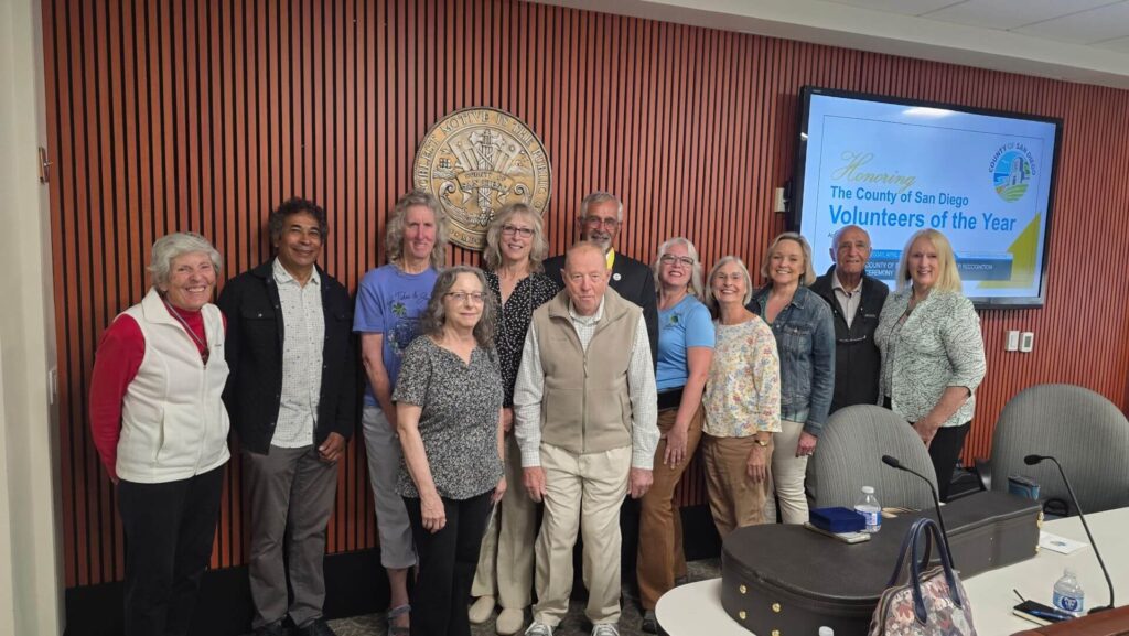 A group of people stand together in a meeting room beside a screen that reads "Volunteers of the Year" and a San Diego County seal on the wall.