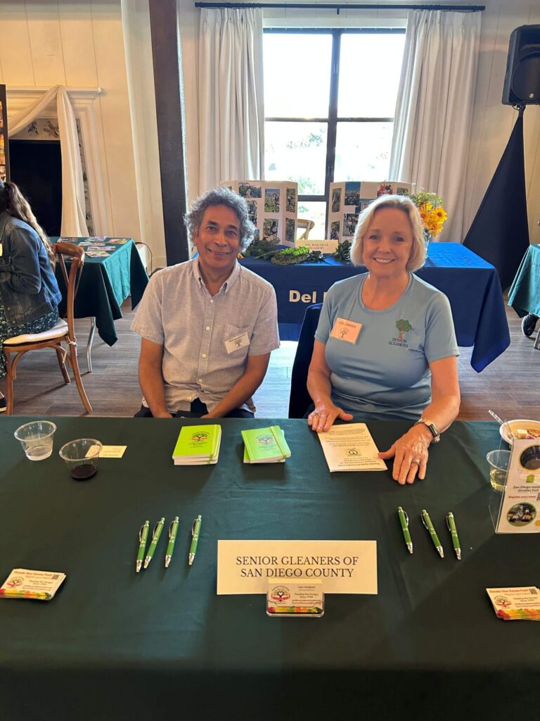 Two smiling individuals behind a table promoting "Senior Gleaners of San Diego County," with informational brochures, pens, and small notebooks displayed.