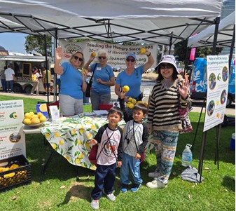 Two children and four adults stand around a community booth under a canopy, with lemons, informational materials, and health education signs displayed on the table.